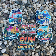 Six colorful stickers featuring California city and scenic designs are arranged on a bed of small gray pebbles. The stickers read Run San Francisco, Run Los Angeles, Run Sacramento, Run San Diego, and Run Modesto, with Run California in the center.