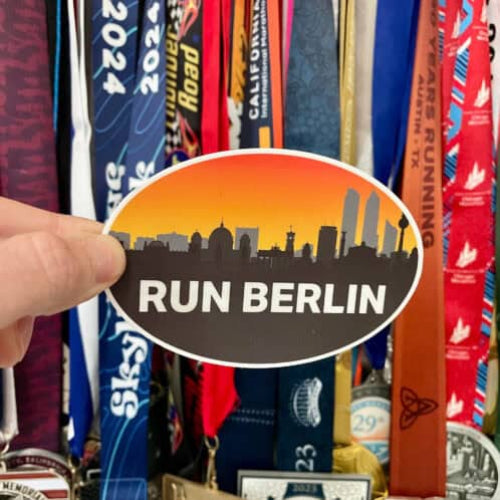 Hand holding a Run Berlin oval sticker in front of a display of race medals and ribbons.