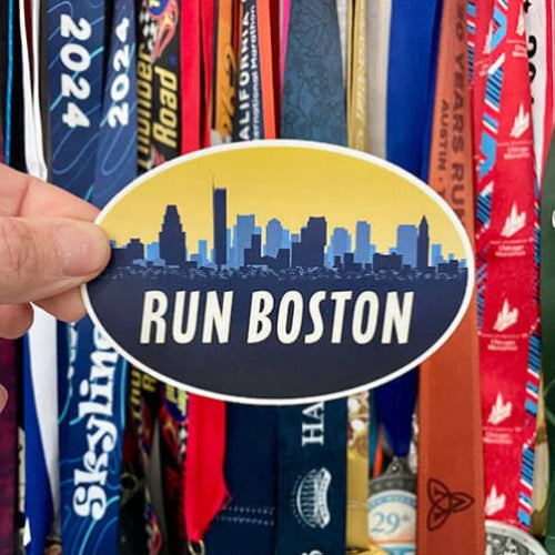 Hand holding a Run Boston oval sticker in front of a display of colorful marathon race bibs and medals.