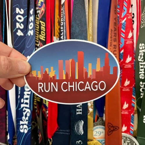 Hand holding a Run Chicago oval sticker in front of a display of race medals and ribbons.