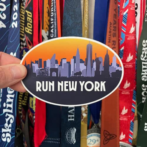 Sticker with 'Run New York' design held over a collection of colorful race medals.