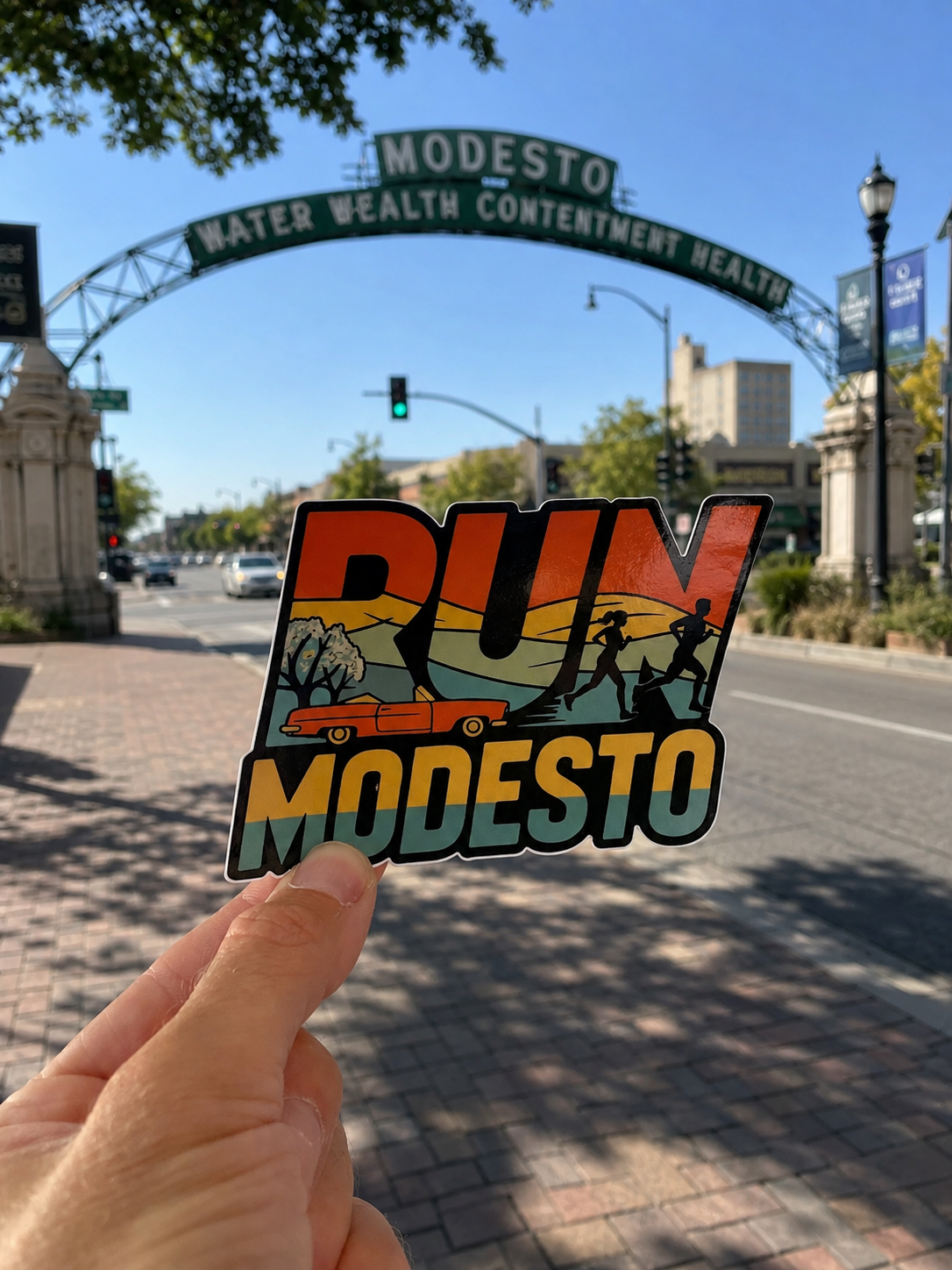 A hand holds a die-cut sticker in front of the Modesto Water Arch in a sunny outdoor city setting. The sticker features the words “RUN MODESTO” with a sunset-style gradient background and silhouettes of a runner, a car, and trees.