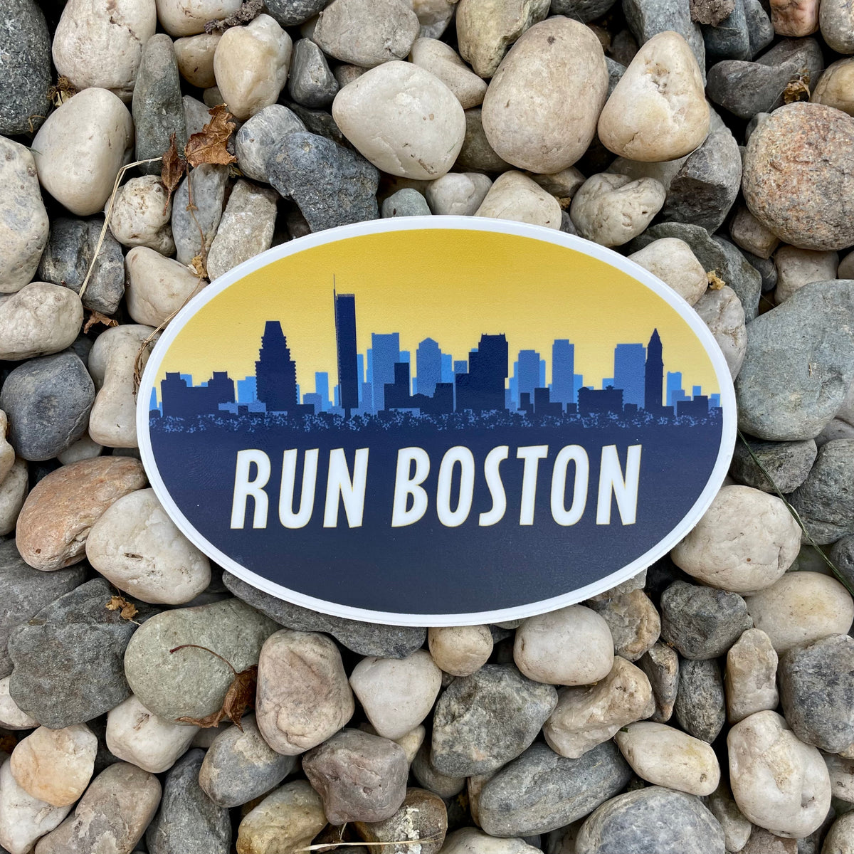 An oval sticker featuring a Boston skyline and the words 'RUN BOSTON' in white, placed on a bed of smooth gray and beige river stones.