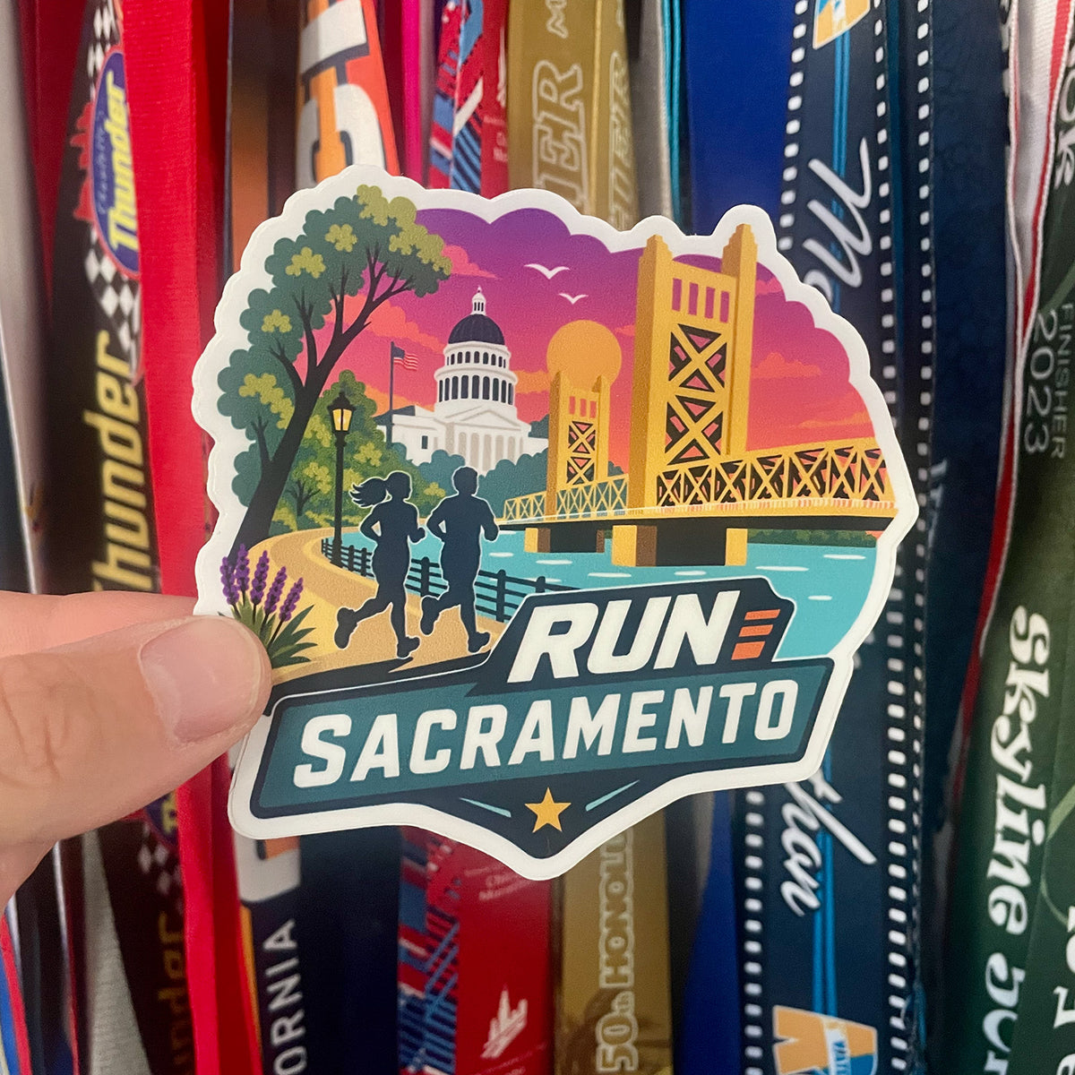 A hand holds a die-cut sticker featuring a colorful illustration of Sacramento landmarks and two runners, with the text 'RUN SACRAMENTO' in large white letters. The sticker is photographed in front of a row of colorful marathon ribbons.
